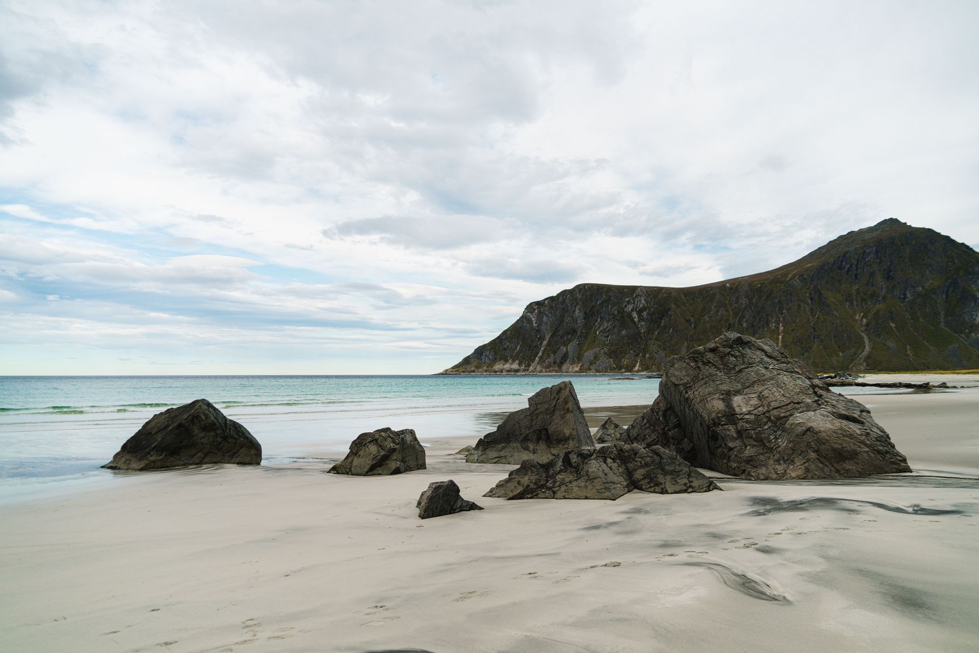 Rocky beach with large boulders, pale sand, calm sea, and a cloudy sky beside a steep green cliff