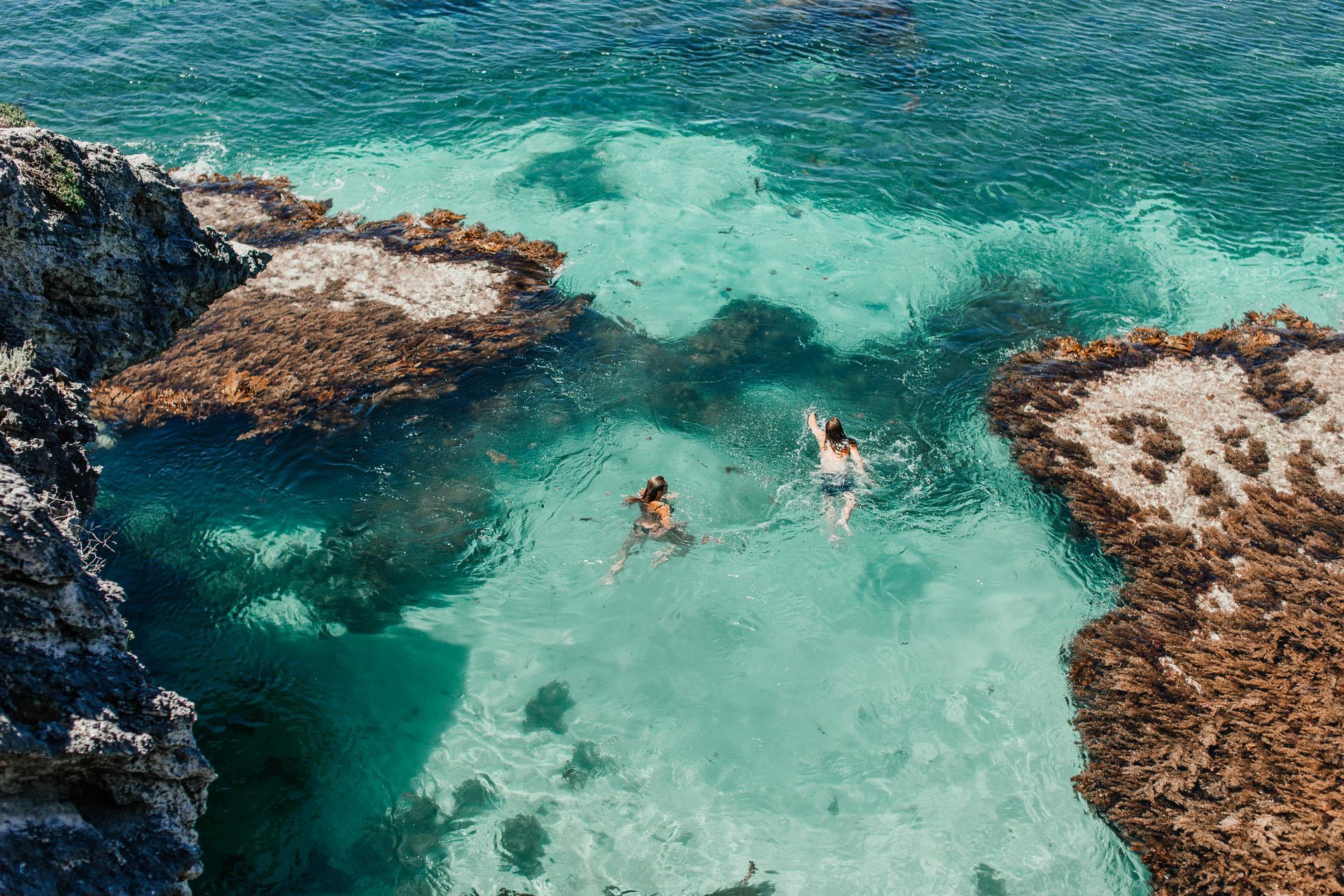 Two swimmers in clear turquoise water between rocky shores and seaweed-covered patches