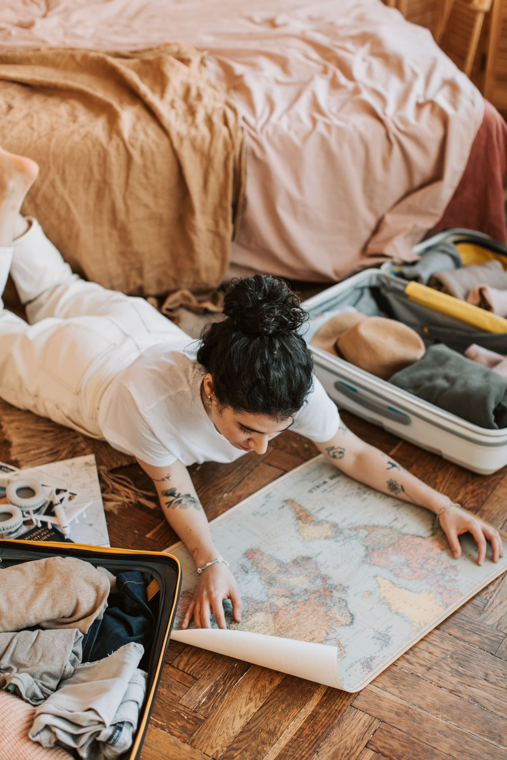 Person kneels on wooden floor, unrolling a map beside an open suitcase in a cozy bedroom.