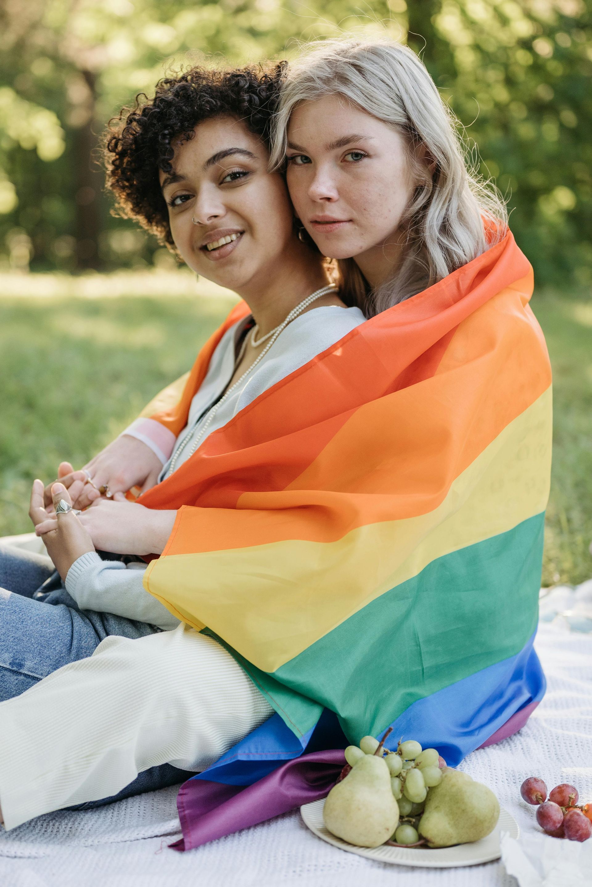 Two people sitting outdoors wrapped in a rainbow pride flag, with fruit on a blanket nearby