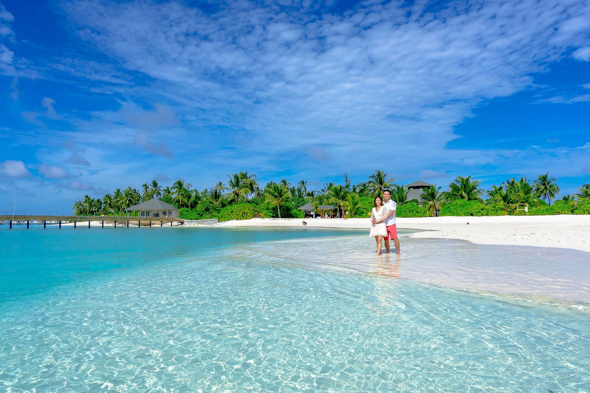 Tropical beach with clear turquoise water, white sand, palm trees, and two people wading near shore