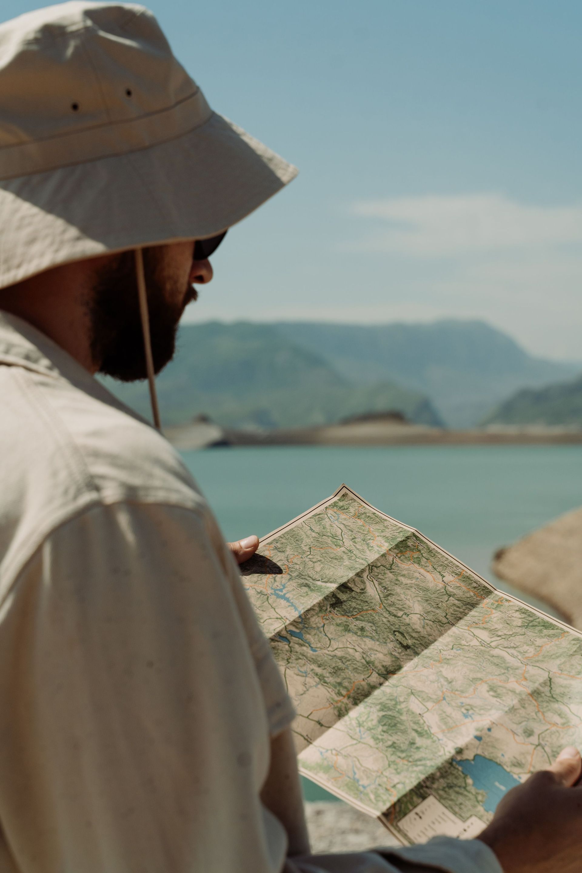 Person in a sun hat holding a map by a turquoise lake with mountains in the background