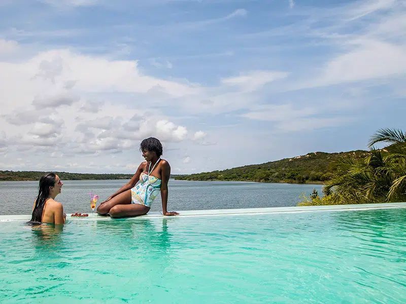 Two people relax at the edge of an infinity pool overlooking a serene lake and forested shoreline under a cloudy sky.