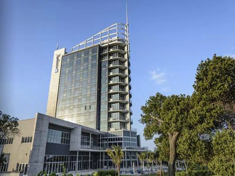Modern glass high-rise building with adjacent low structure and trees under a clear blue sky