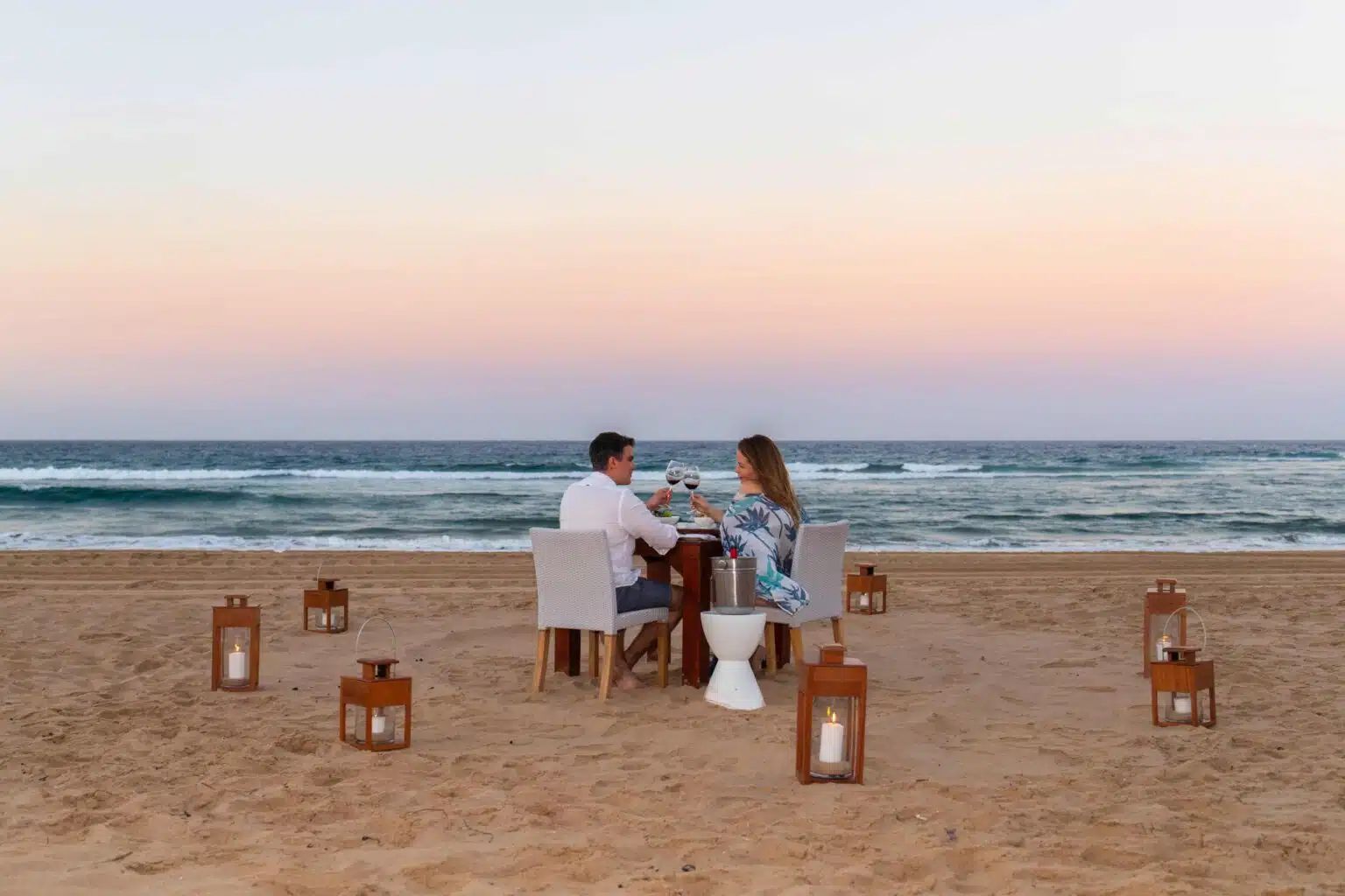 A couple having a romantic dinner at a table on a sandy beach at sunset, surrounded by lit lanterns.