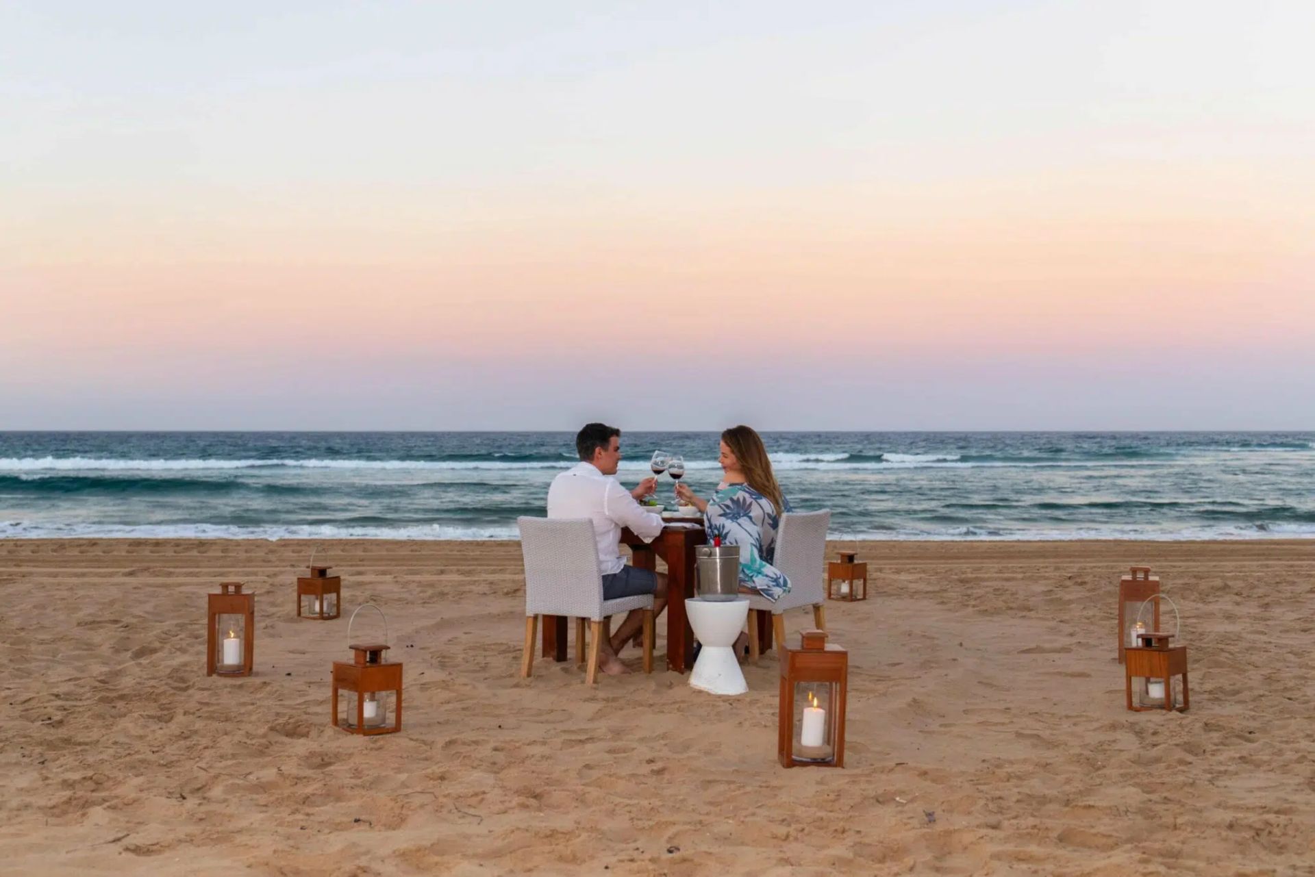 Couple dining at a beach table at sunset, with lanterns on the sand and ocean waves behind them