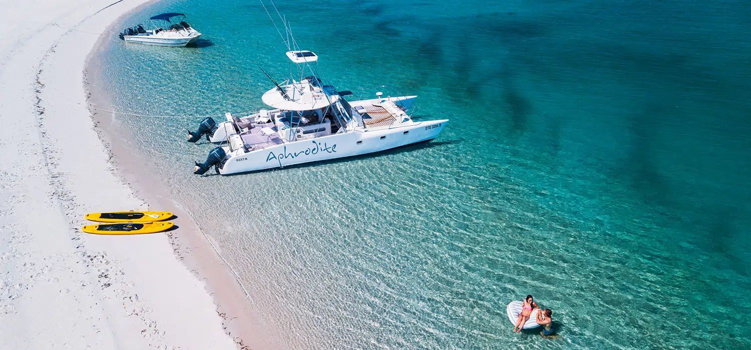 Aerial view of a sandy beach with turquoise water, a white yacht, smaller boats, and a yellow kayak