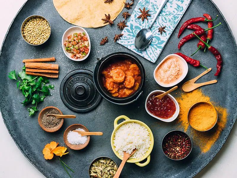 Assorted spices, herbs, and sauces arranged around a black pot on a textured blue tabletop.