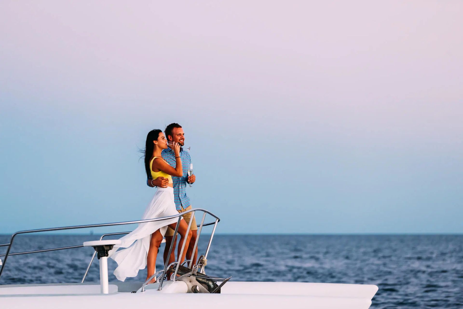 Couple standing on a sailboat deck at sea during sunset, embracing and looking out over the water
