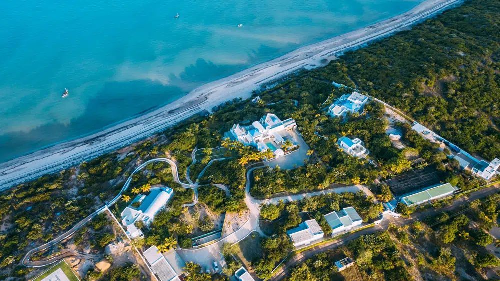 Aerial view of a tropical beach resort with white buildings, palm trees, and turquoise ocean shoreline.