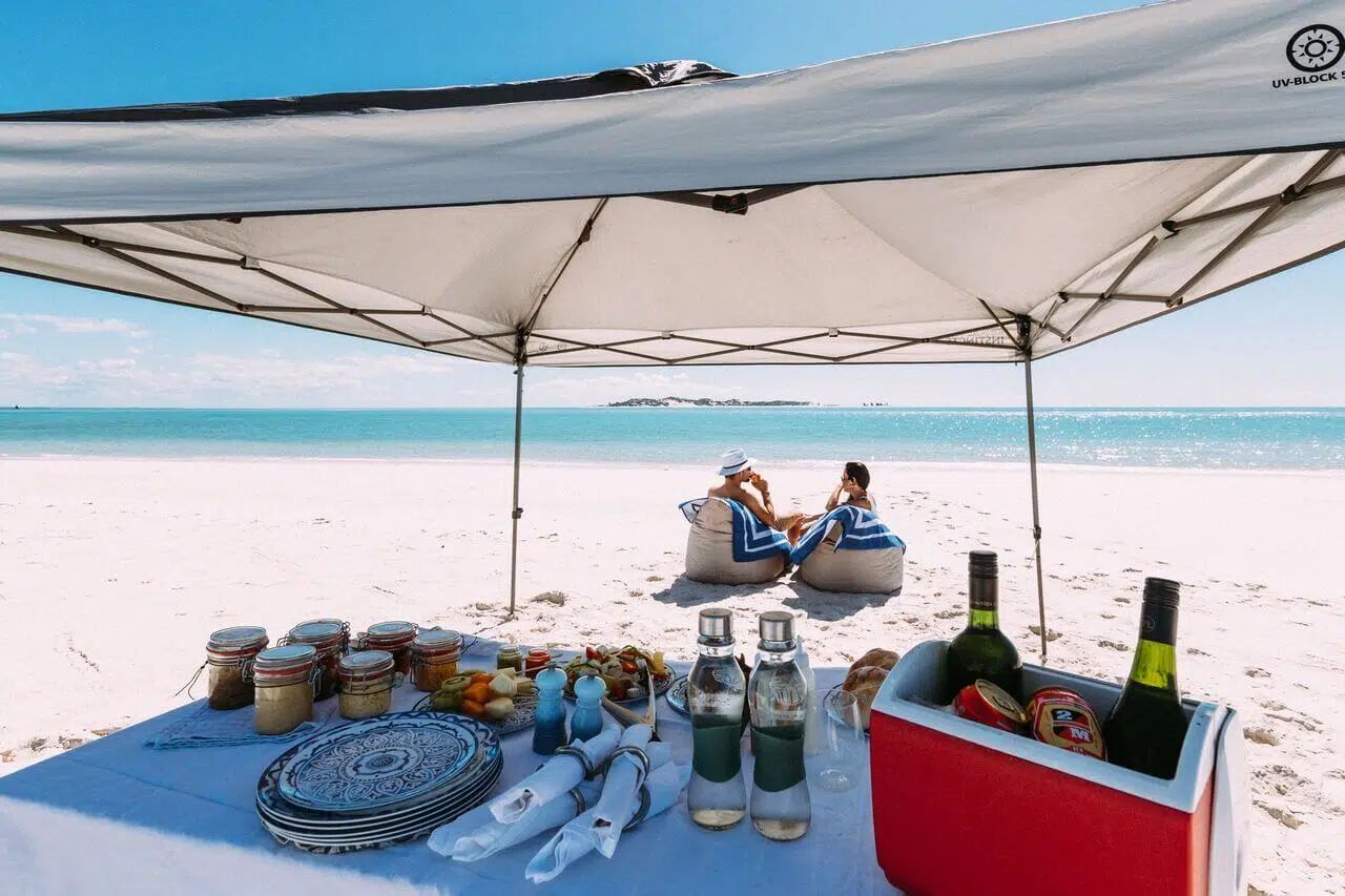 Beach picnic under a white canopy with two people sitting by the turquoise ocean, drinks and snacks in front of them