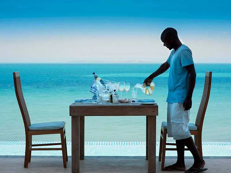 Man standing by a table set for drinks on a beach, with turquoise ocean in the background