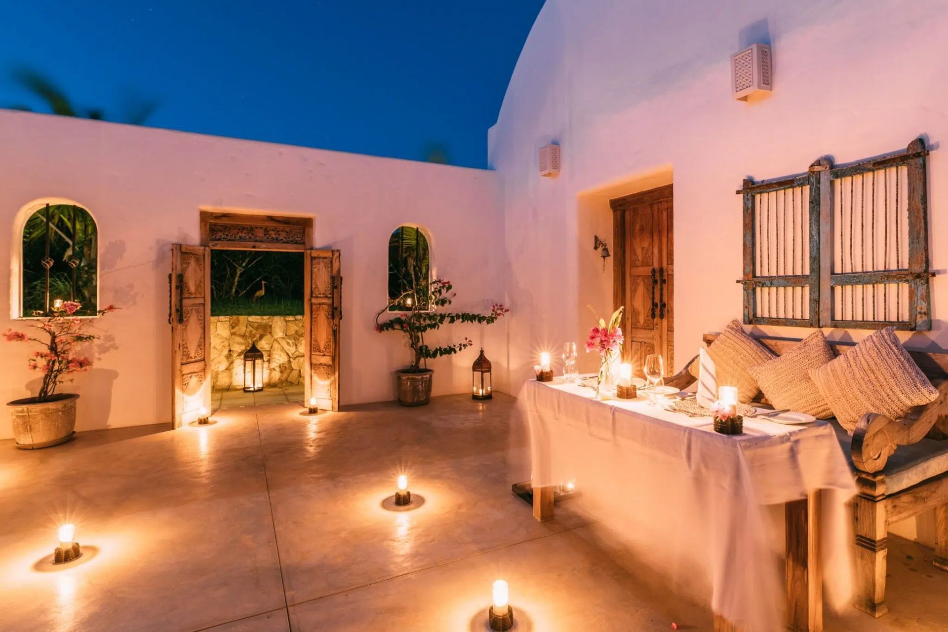 Candlelit rooftop patio with white walls, low table, cushions, and lanterns glowing at dusk