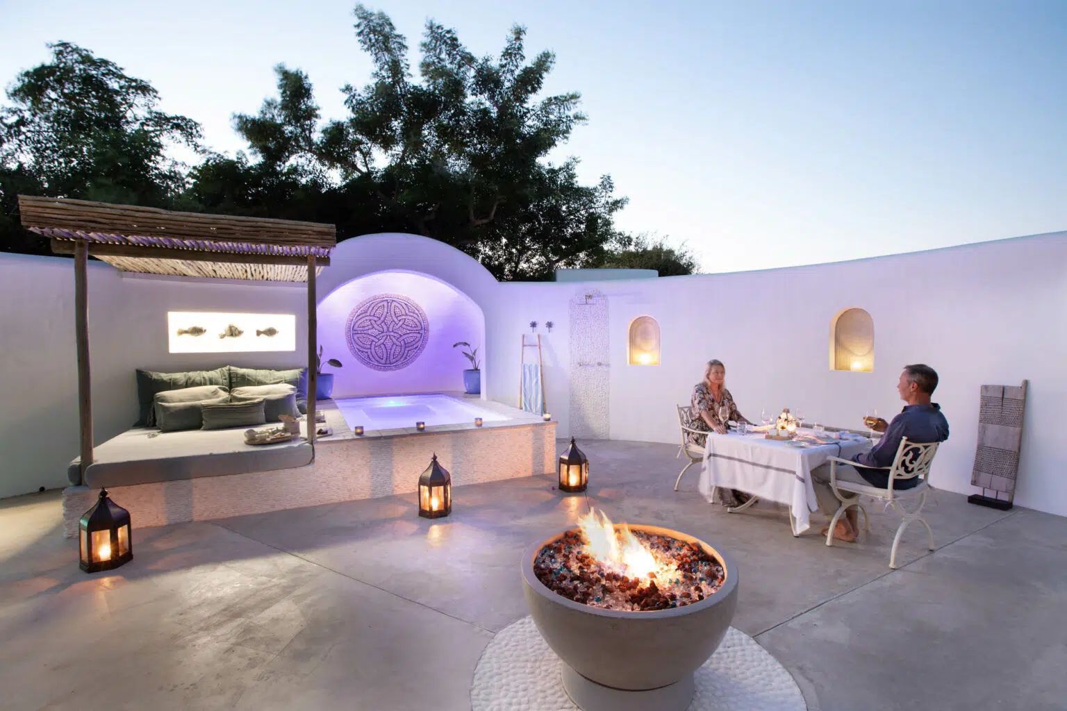 A couple dines at a table on a patio featuring a fire pit, a covered daybed, and a swimming pool against a white wall.