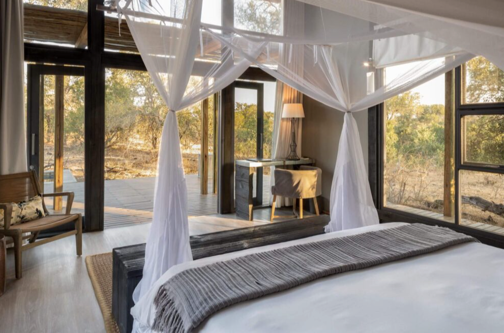 Bright bedroom with a canopy bed, sheer white drapes, and large windows overlooking autumn trees