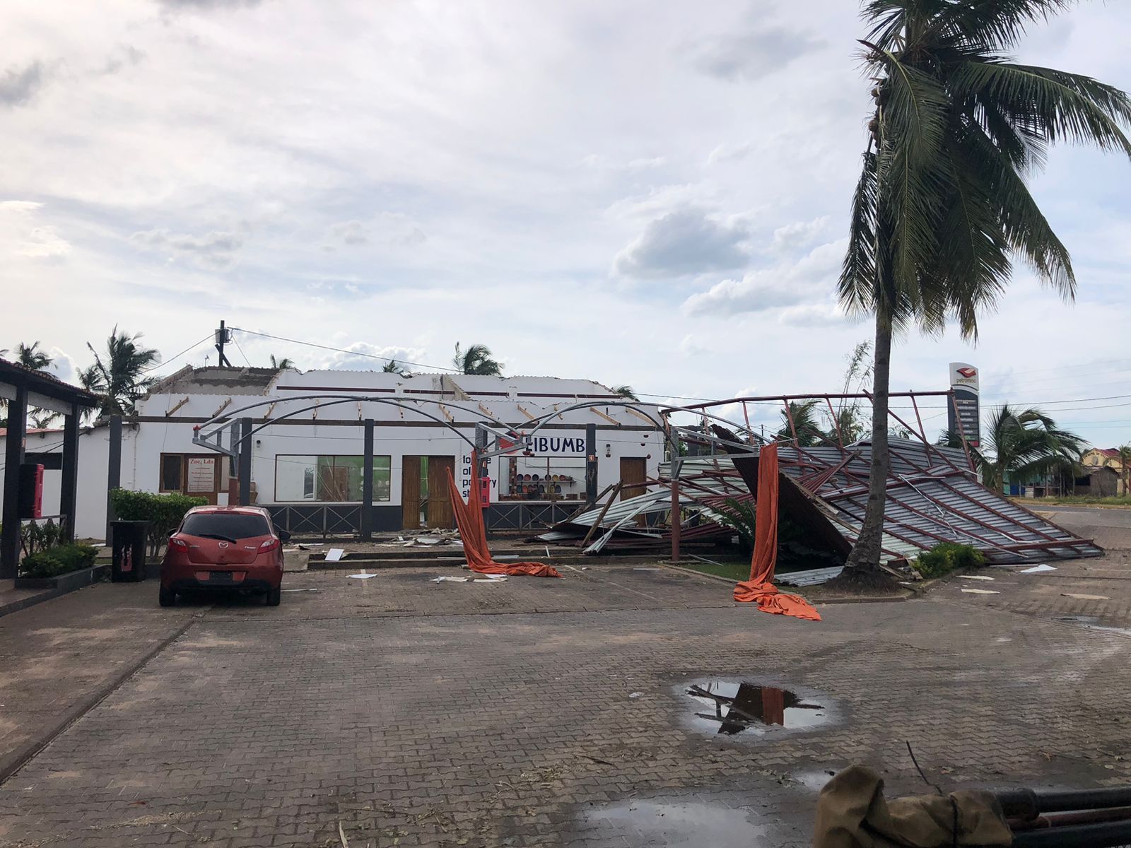 Collapsed roof and damaged building with debris, a red car, and a palm tree in the foreground.