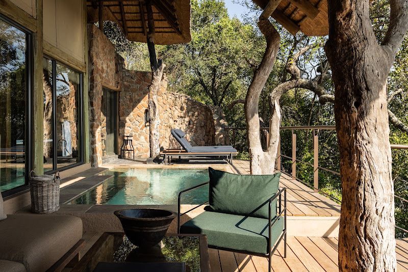 Modern deck with green seating, glass walls, and a forest view beside a rustic stone cabin