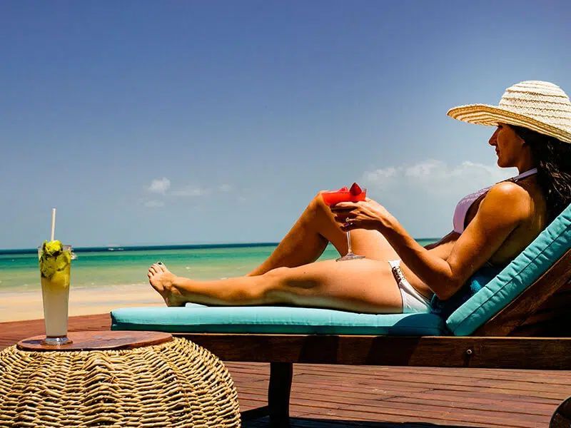 Woman relaxing on a lounge chair by the beach, wearing a sunhat and holding a drink under a clear sky.