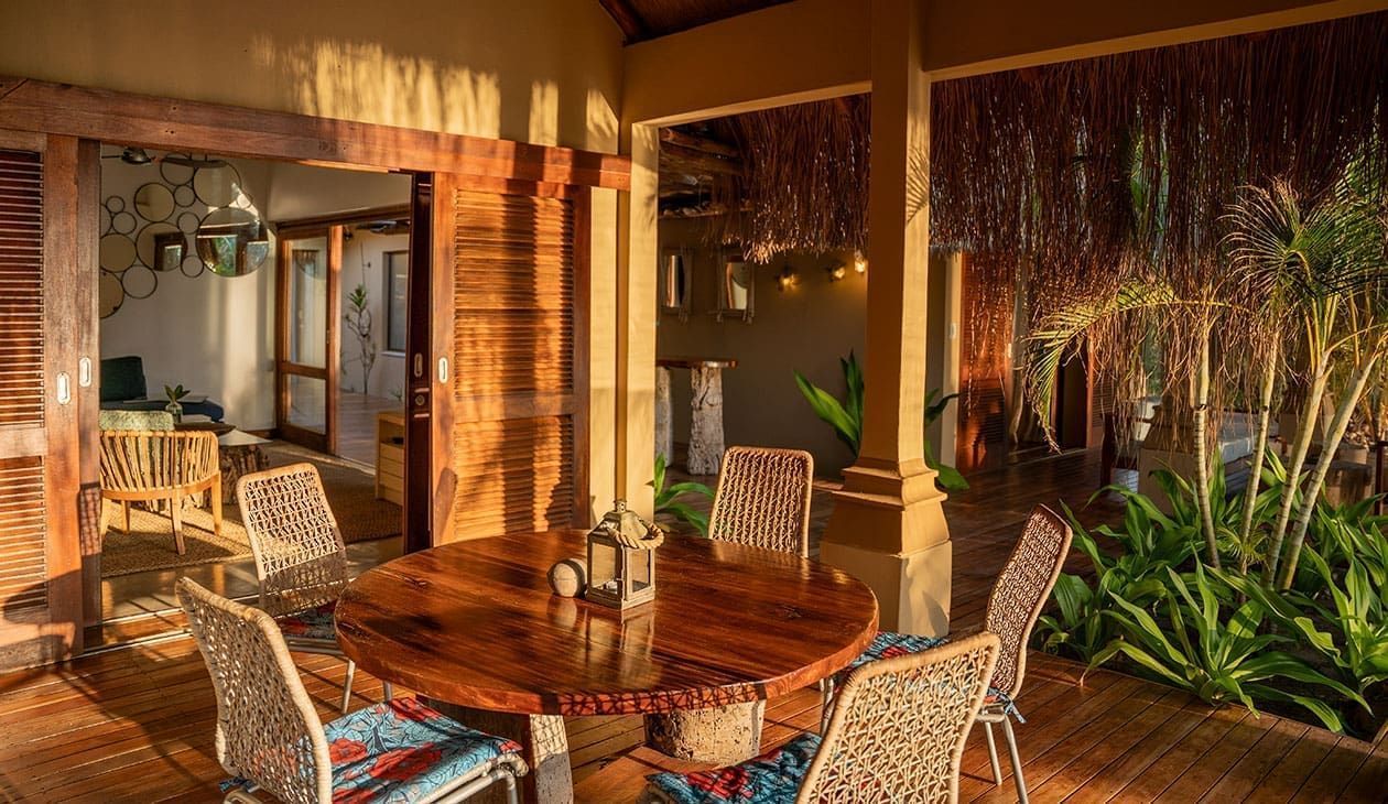 Sunlit patio with wicker dining chairs, wooden table, and tropical plants beside a home.