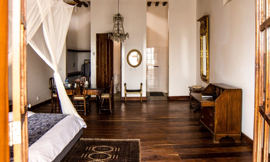 Bright, airy bedroom with a four-poster bed, wooden floor, round mirror, and simple antique furniture