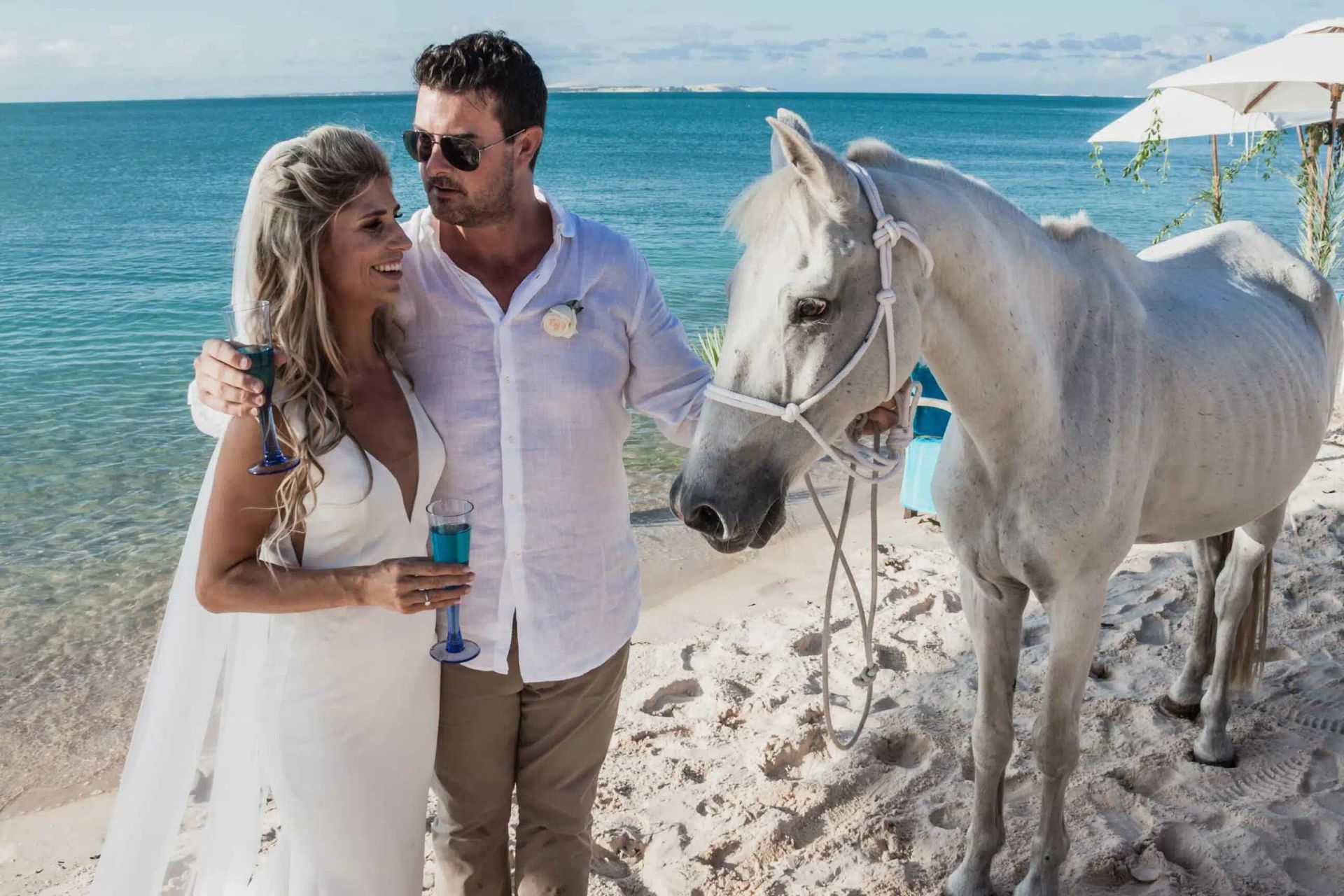 Couple standing on a sunny beach beside a white horse near the turquoise sea