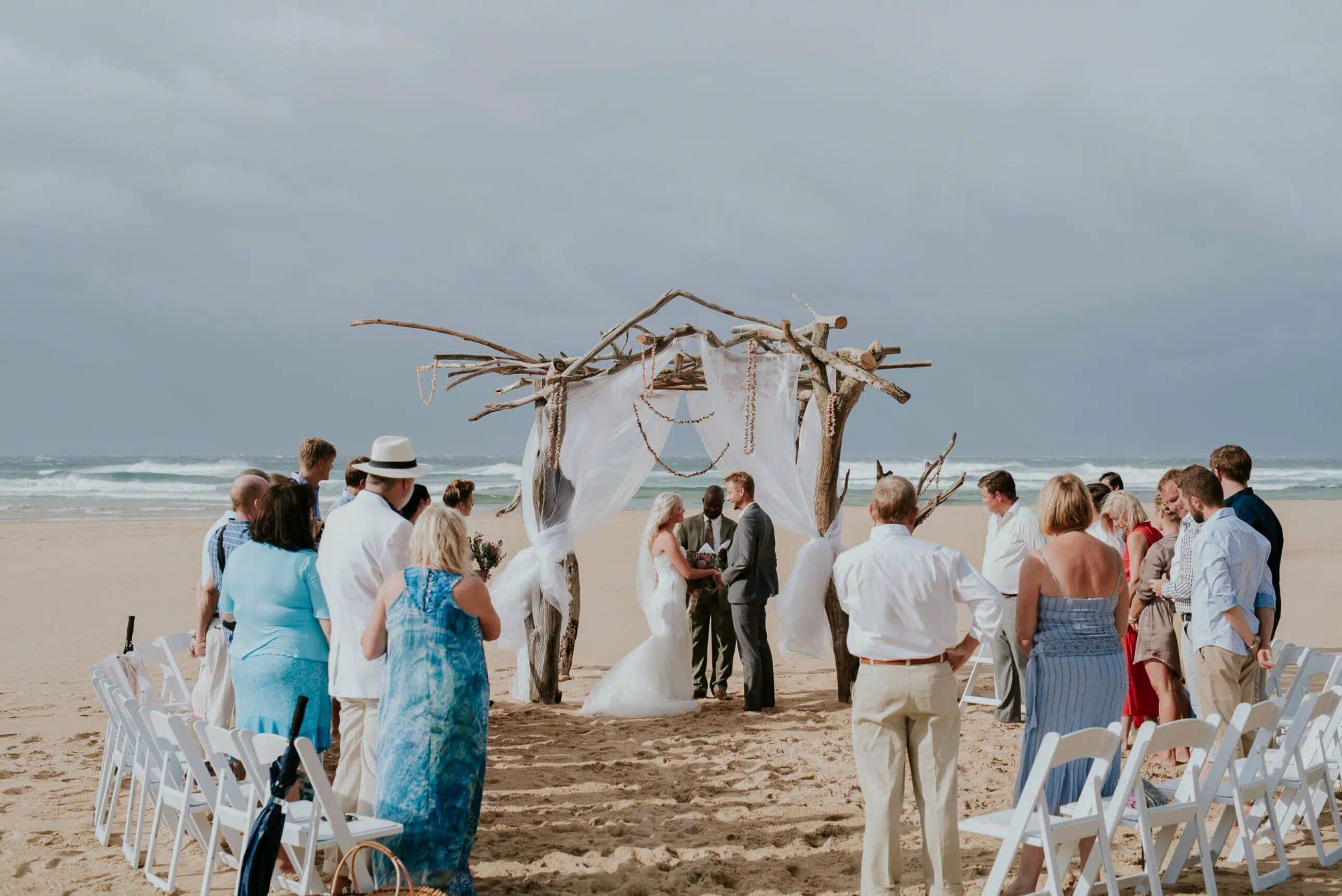 Beach wedding ceremony under a driftwood arch with guests gathered around in white chairs
