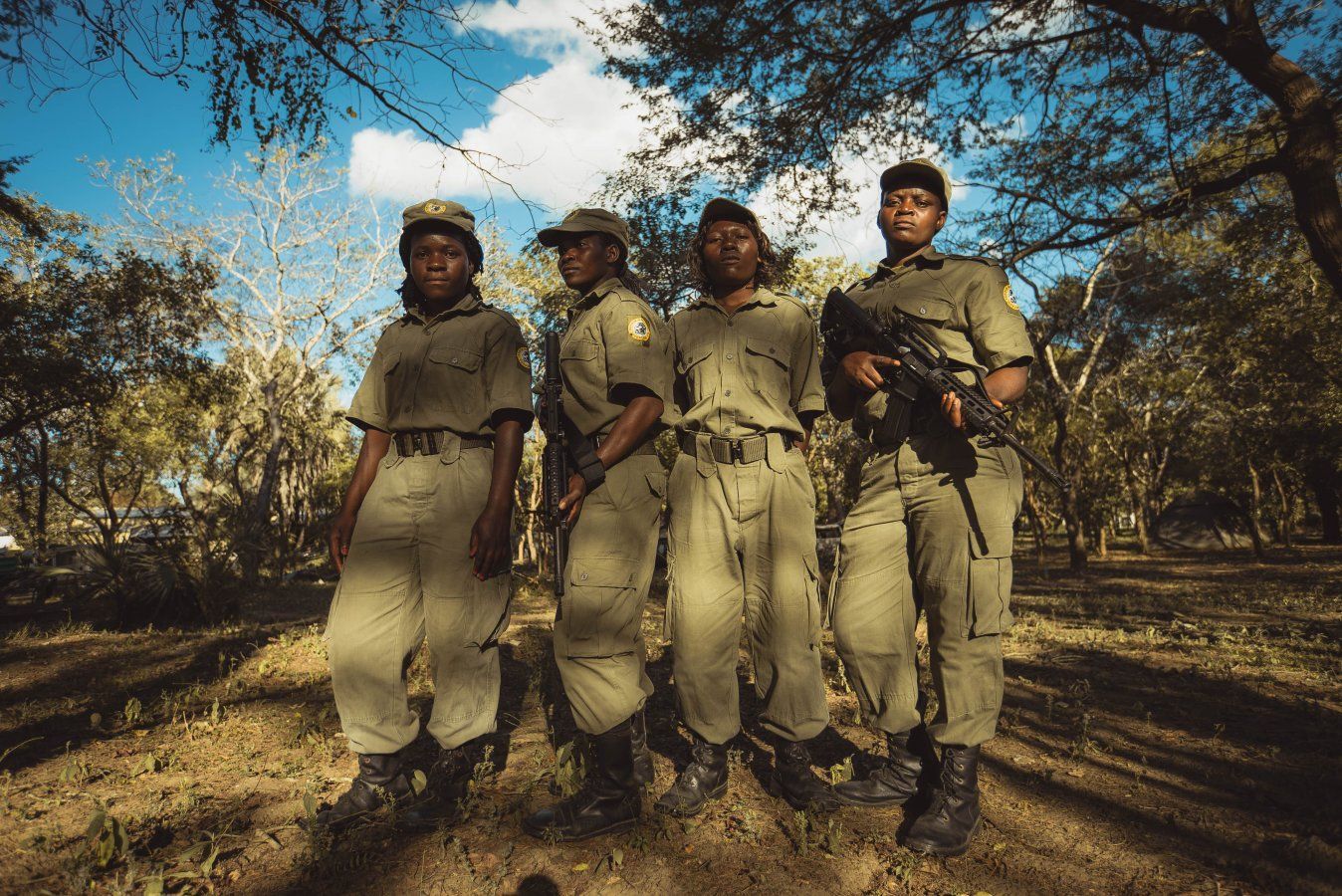 Four soldiers in tan uniforms walk along a wooded path, rifles in hand, under bright sunlight.