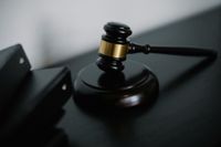 A wooden gavel with a gold band rests on its sound block beside a stack of black binders on a dark surface.