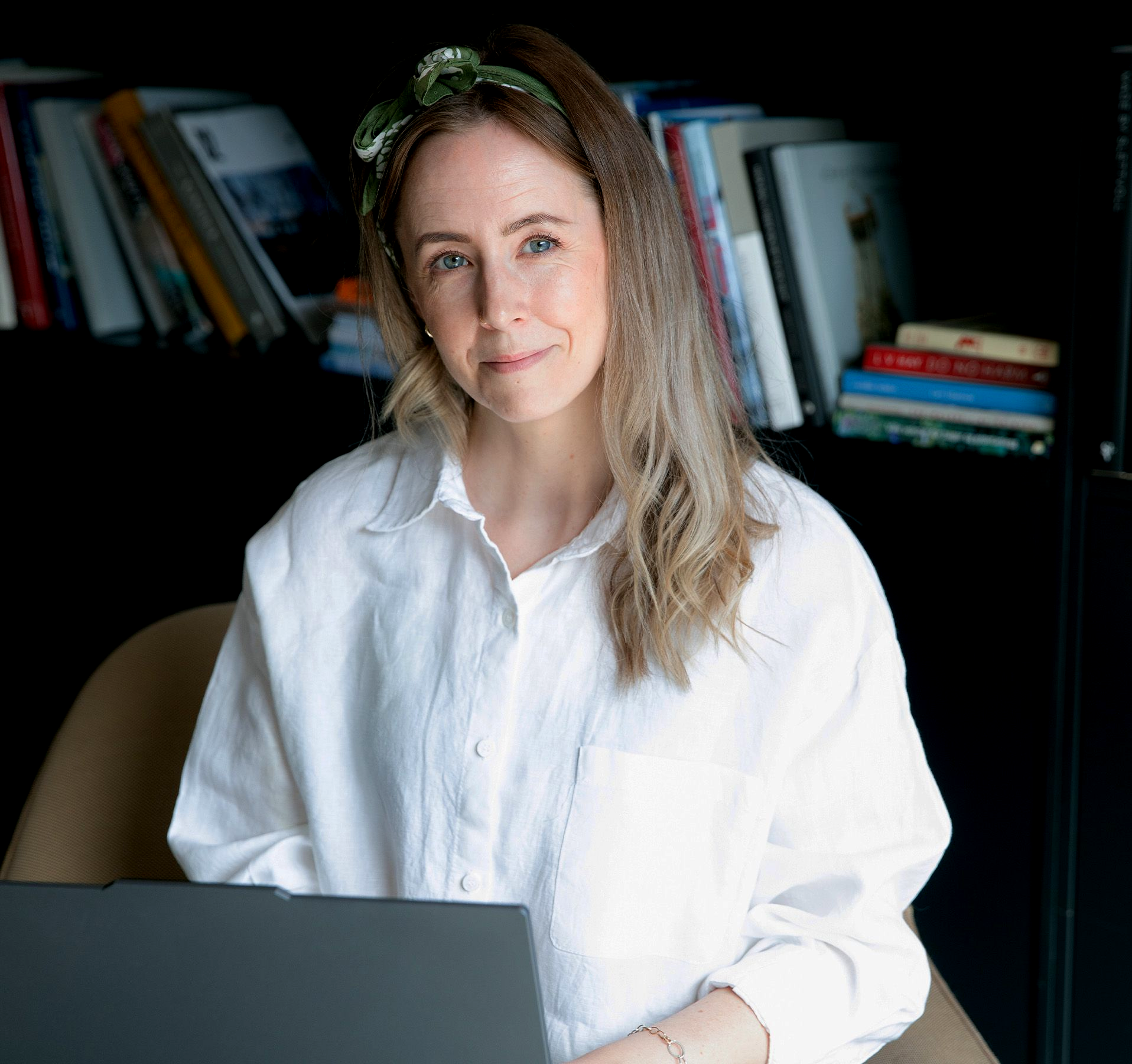 Monica Myklebust med hvit skjorte og laptop på fanget. Smiler til kamera.