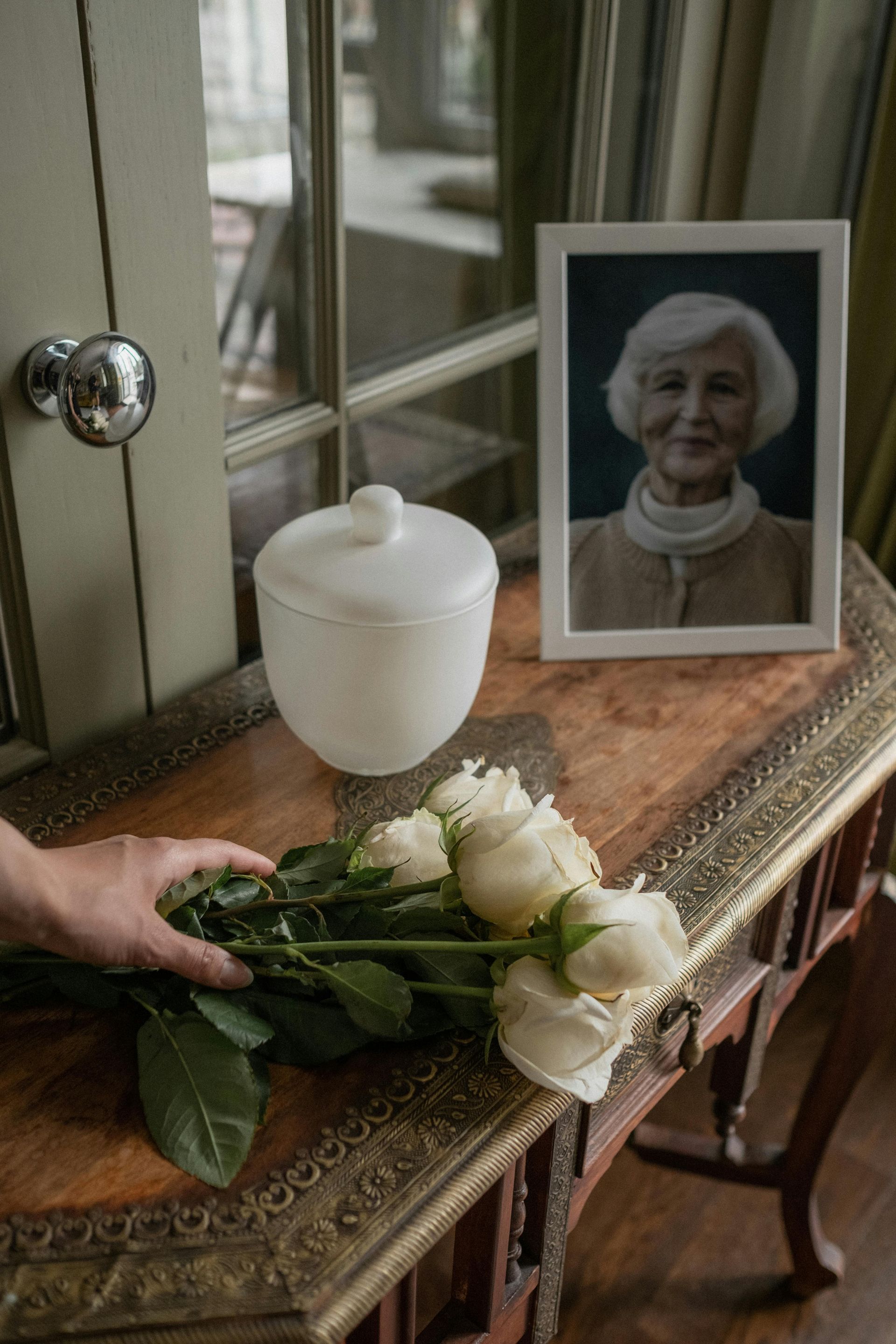 A hand placing white roses on a table next to a portrait and urn.