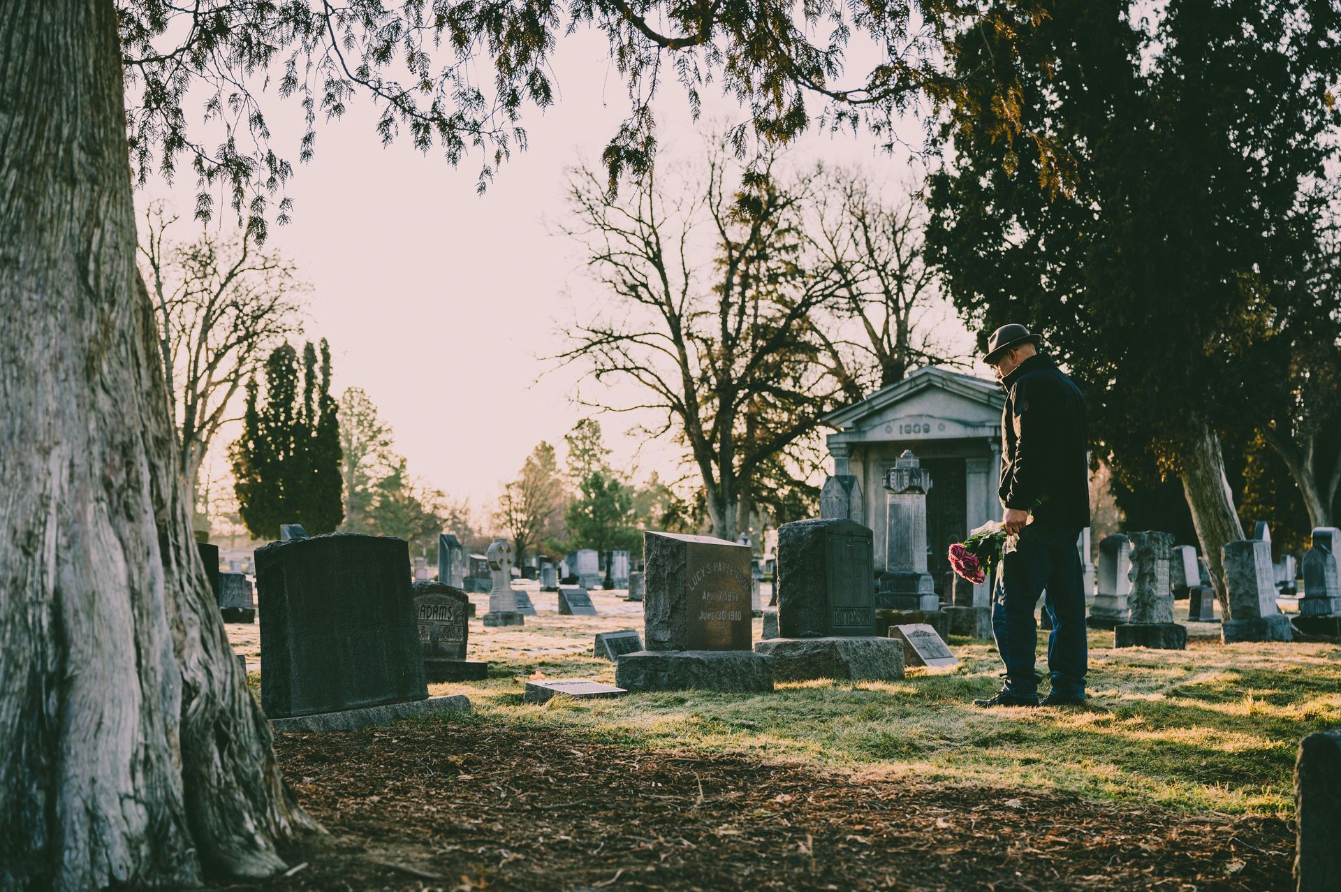 Person standing in a graveyard, holding flowers near a headstone at sunset.