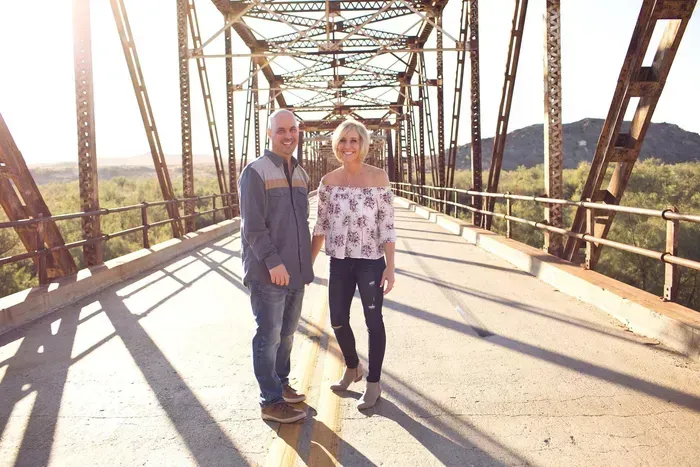 Two people standing on a sunlit metal bridge with hills in the background