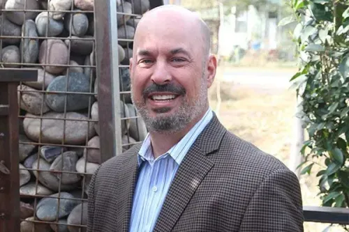 Smiling bald man in a checkered blazer standing outdoors beside a stone wall