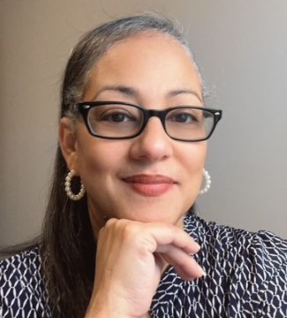 A woman wearing glasses and hoop earrings is looking at the camera.