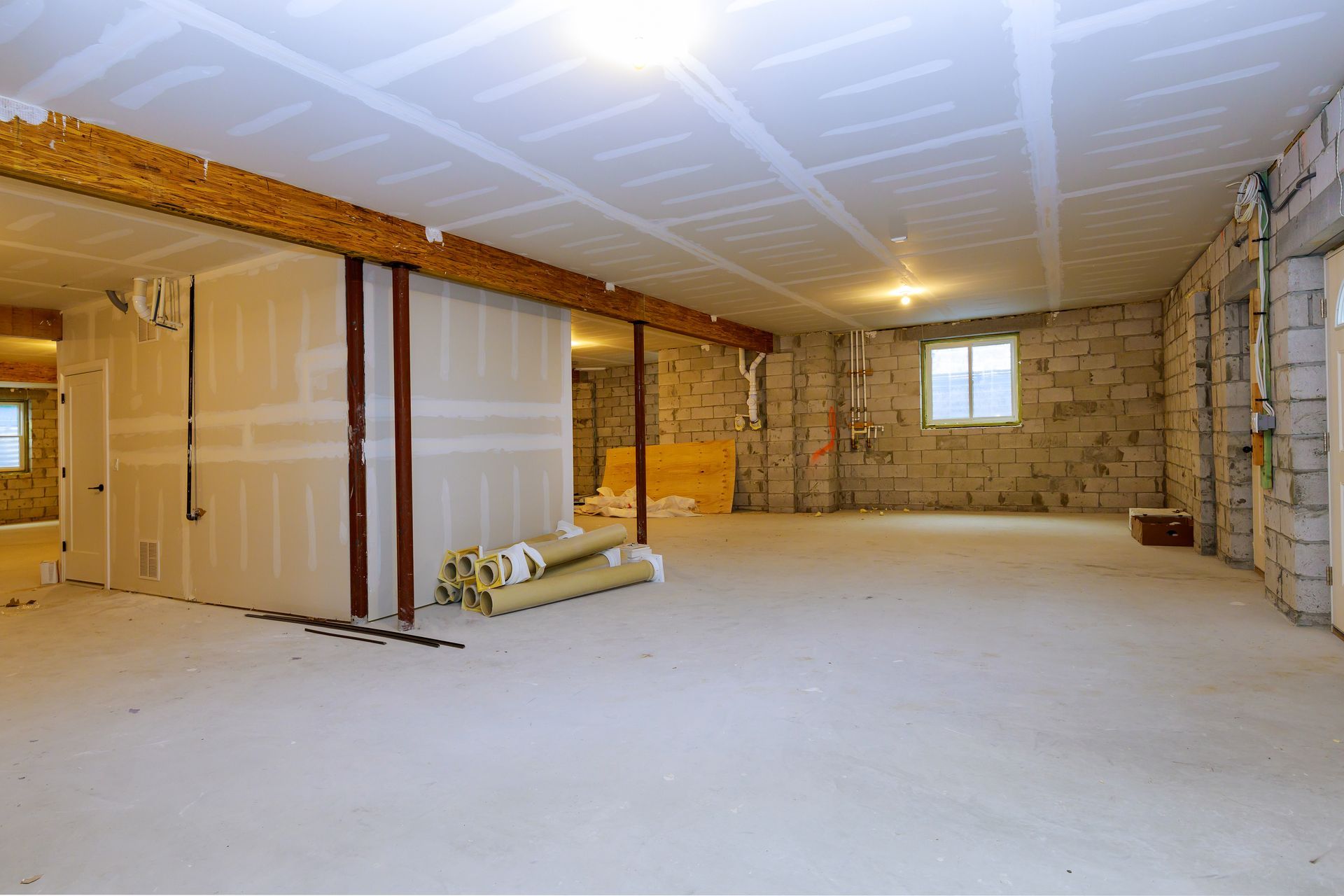 A basement interior under construction with drywall, exposed concrete blocks, a wooden support beam, and rolled flooring. A basement interior under construction with drywall, exposed concrete blocks, a wooden support beam, and rolled flooring.