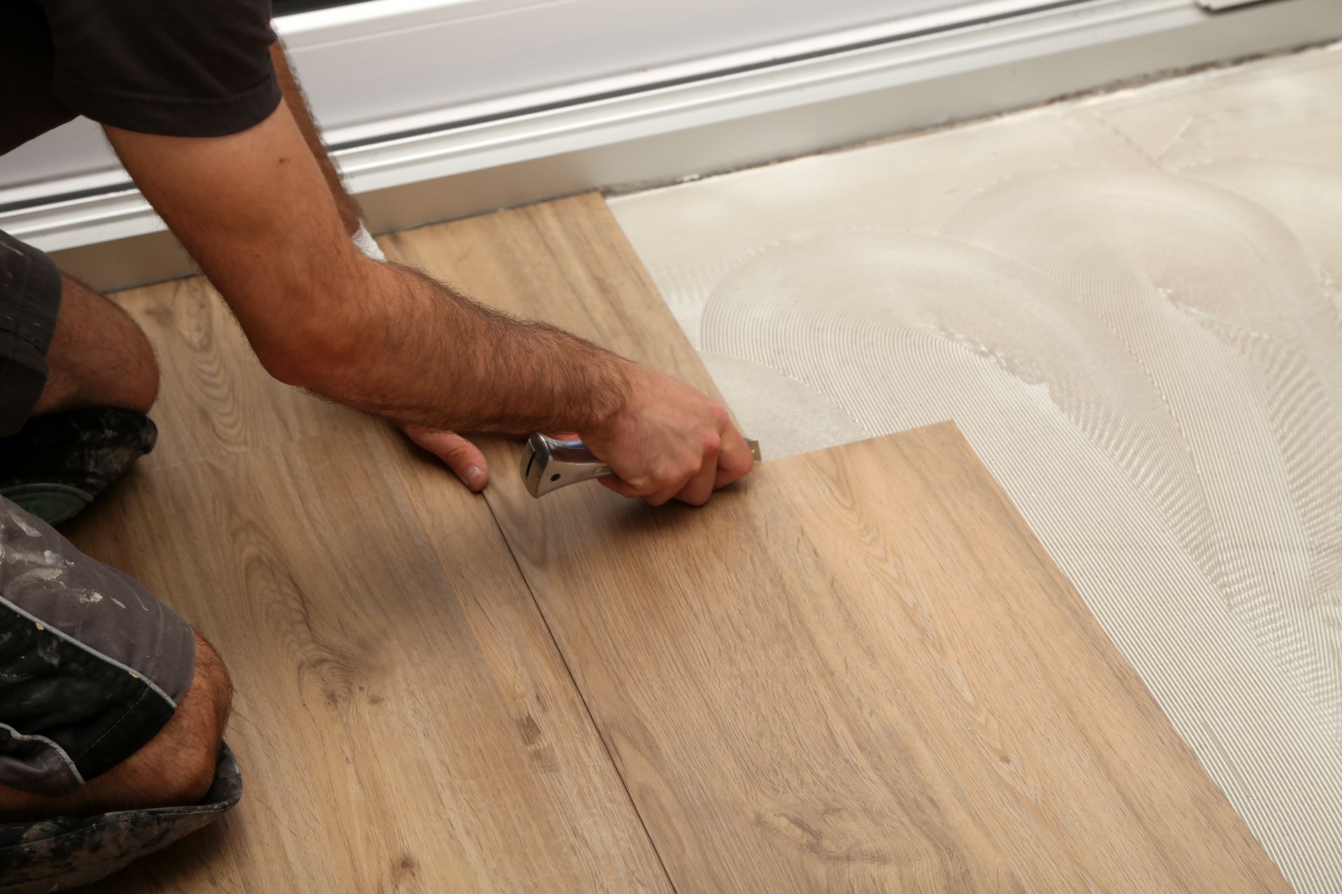 A worker installs wood-look flooring planks over an adhesive-covered subfloor next to a wall baseboard. A worker installs wood-look flooring planks over an adhesive-covered subfloor next to a wall baseboard.