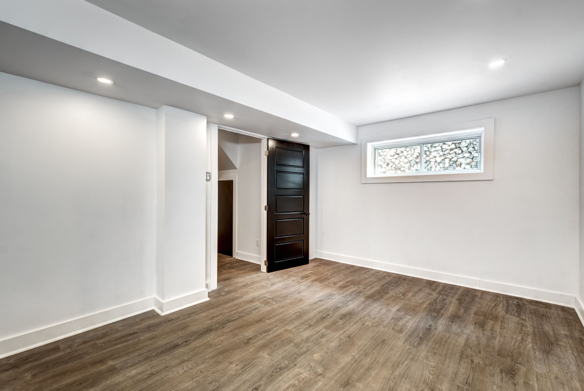 A bright, empty basement room with white walls, wood-look flooring, recessed lighting, and a dark door. A bright, empty basement room with white walls, wood-look flooring, recessed lighting, and a dark door.