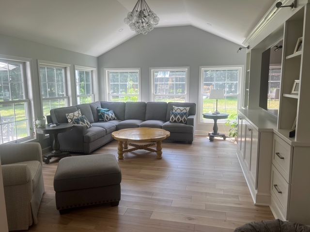 A sunlit living room with a large gray sectional sofa, round wooden coffee table, ottoman, and built-in white cabinets.