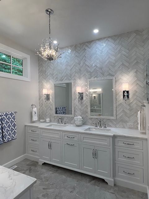 A double vanity bathroom with white cabinets, marble herringbone tile wall, two mirrors, sconces, and a crystal chandelier.