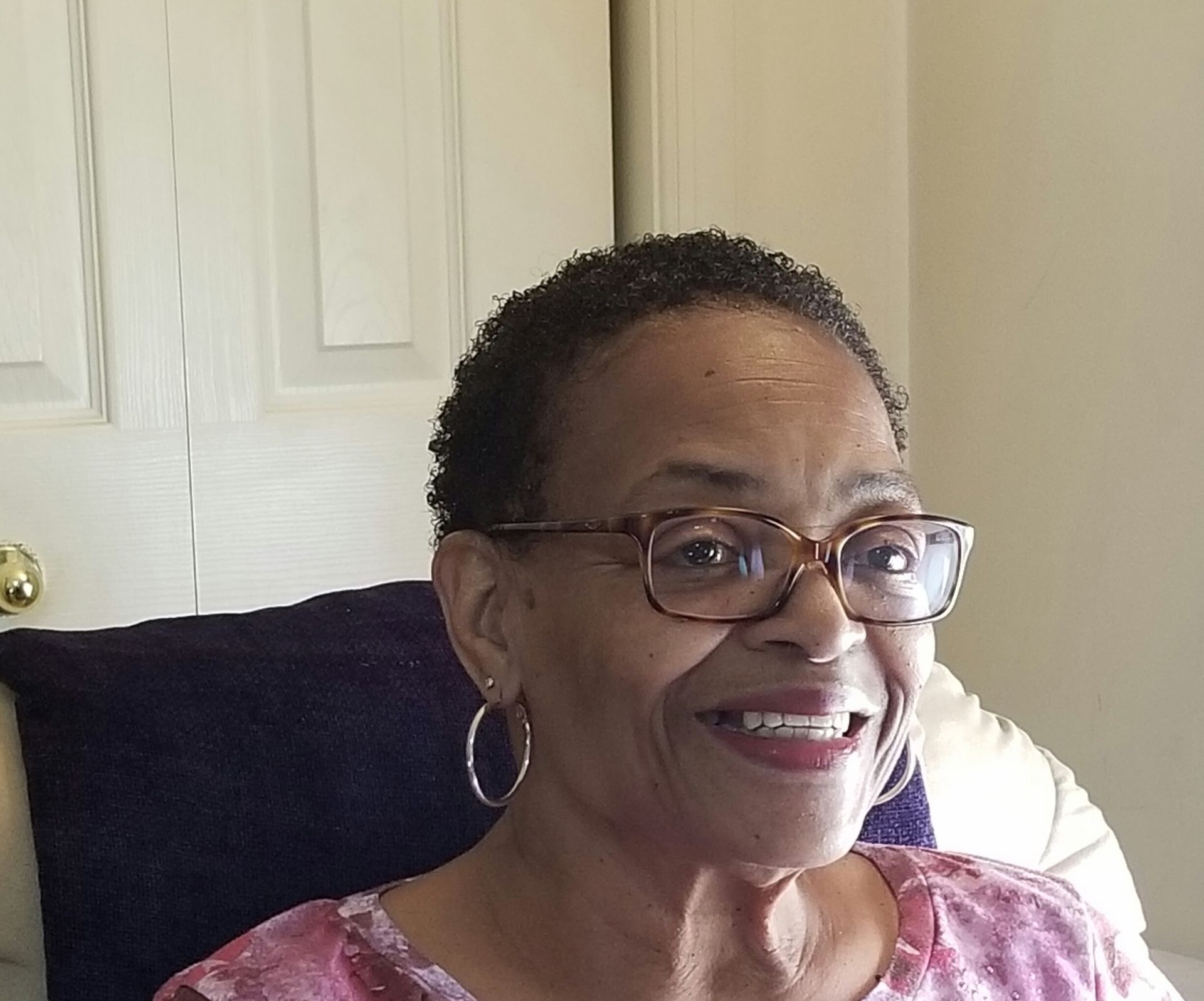 Smiling woman wearing glasses and hoop earrings, seated indoors against a light wall.
