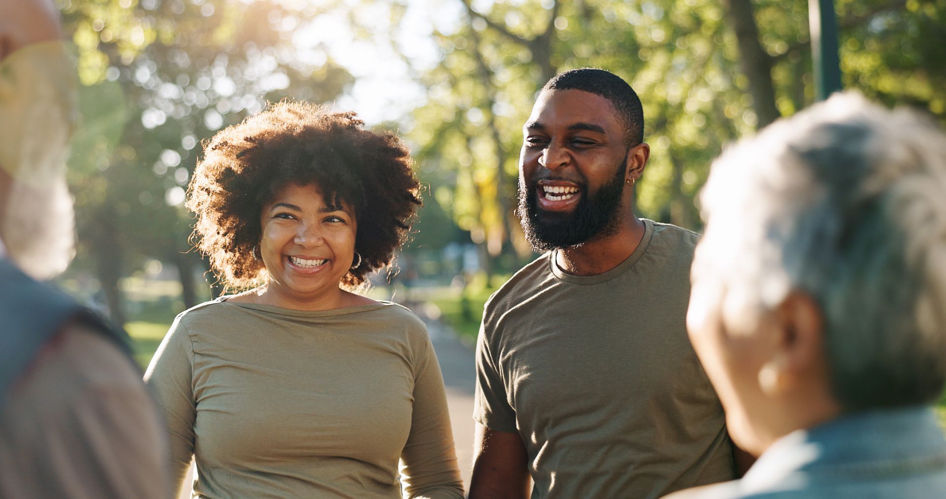 Three smiling people chatting outdoors in sunlight, with trees blurred in the background.