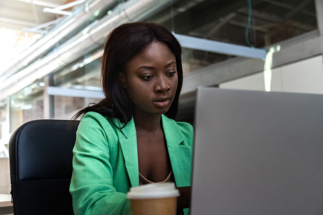 Woman in a green blazer working on a laptop in an office, with a takeaway coffee cup nearby.