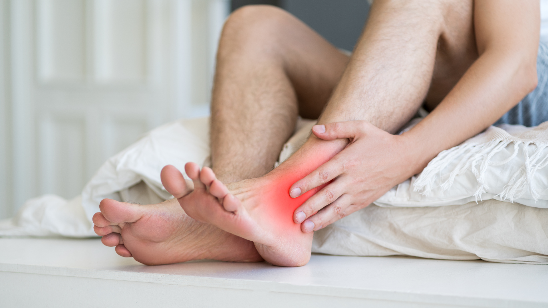 Person sitting on a bed, holding ankle that shows red inflammation.