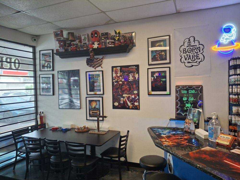 Small vape shop interior with tables, stools, wall art, neon signs, and a counter display.