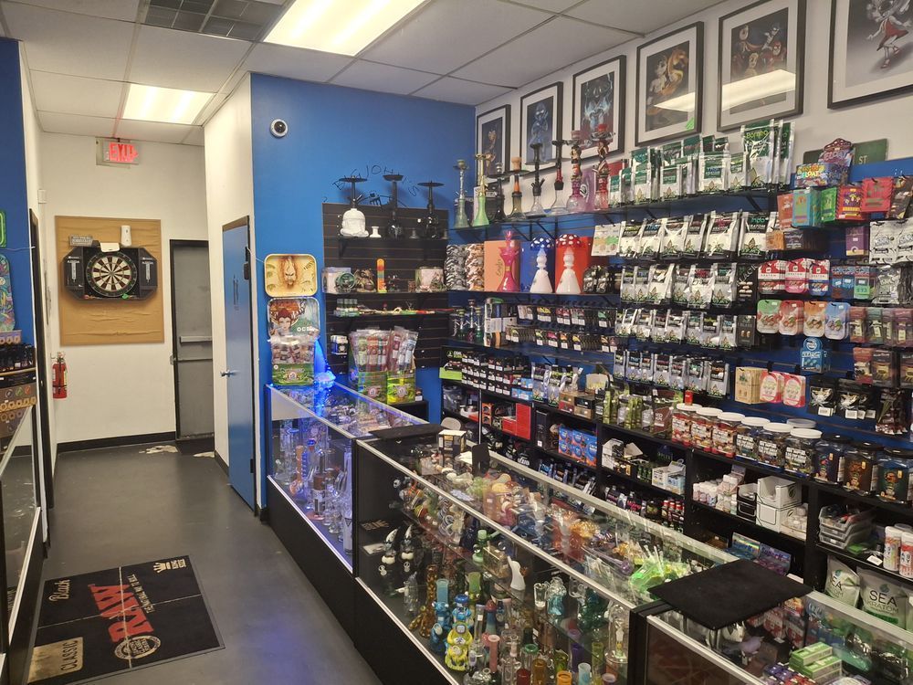 Convenience store aisle with shelves of snacks, drinks, and a dartboard by the door