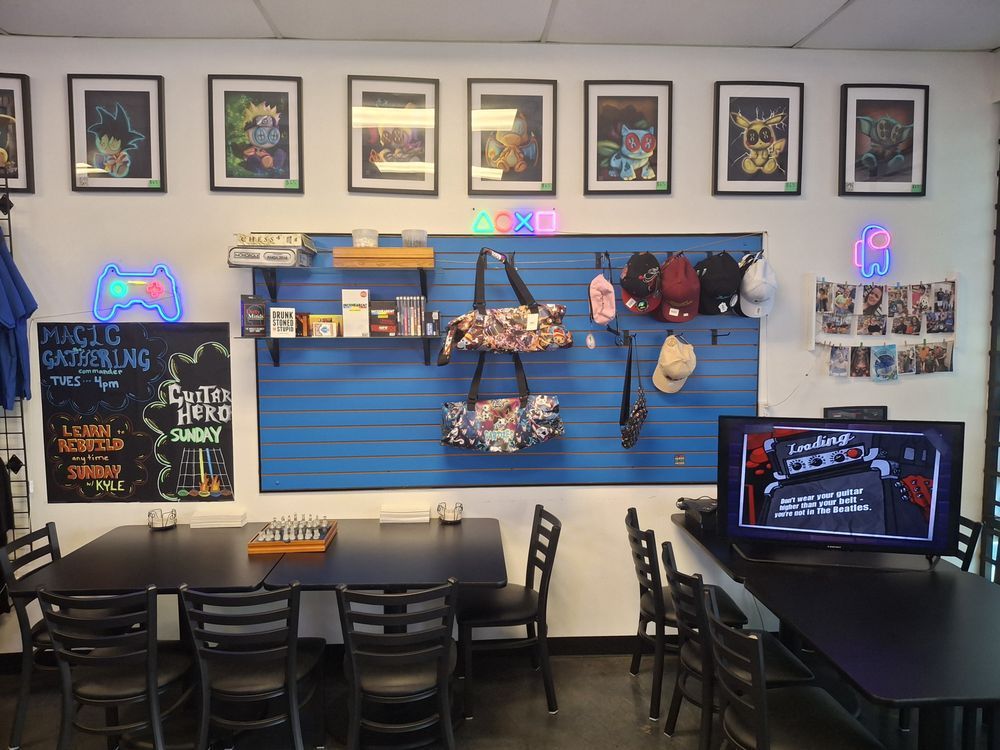 Small arcade/game room with blue wall display, framed art, tables, chairs, and neon signs.