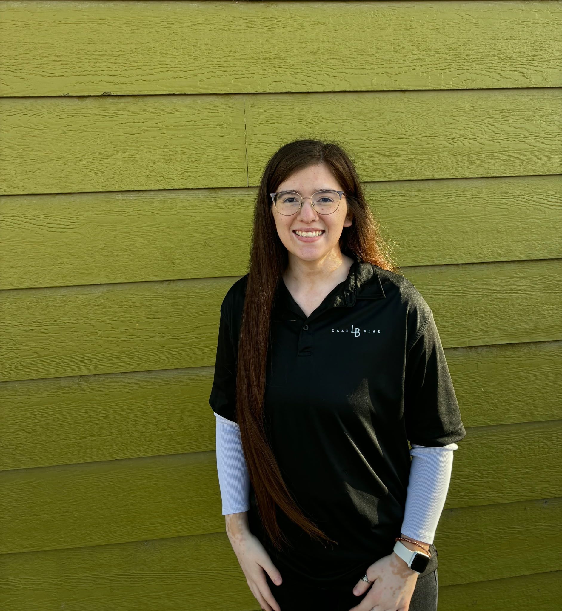 Woman in black shirt and white sleeves standing against a green textured wall, smiling.