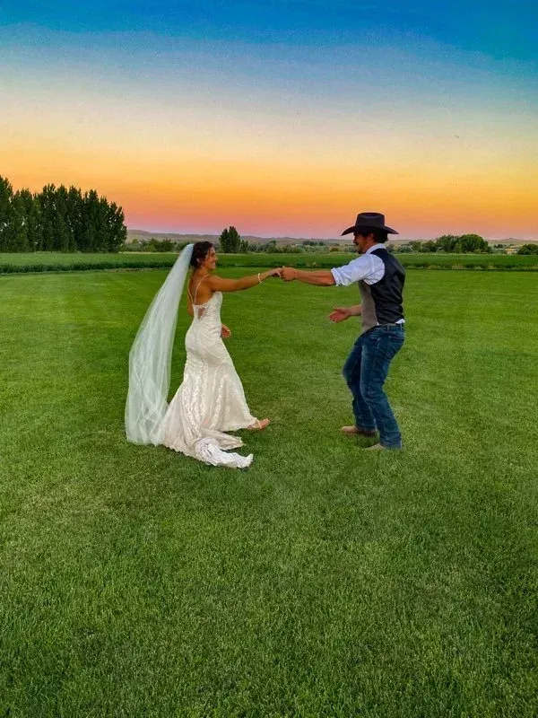Bride and cowboy holding hands in a green field at sunset under a colorful sky