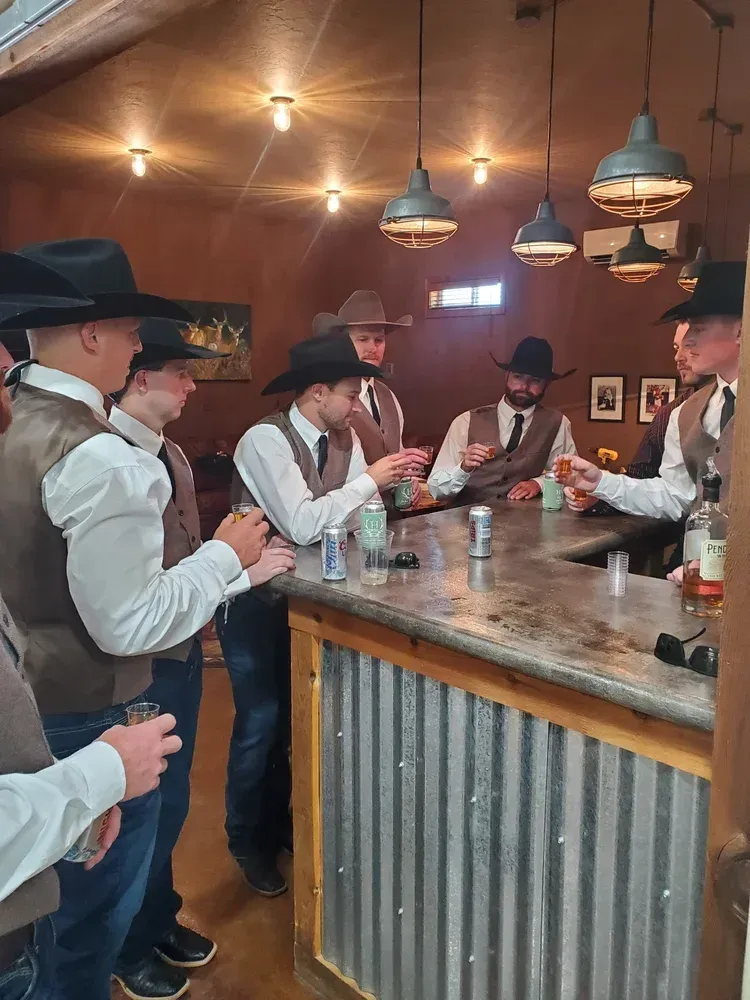 Cowboys in hats and vests socializing at a rustic wooden bar with hanging lights