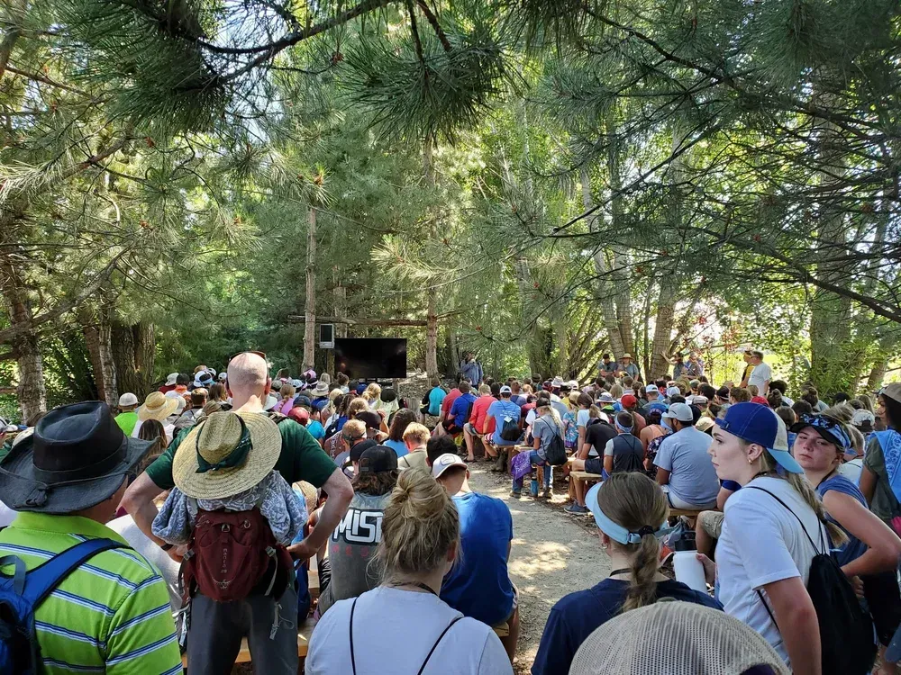 Crowd gathered on a wooded trail under tall trees, some wearing hats and backpacks.